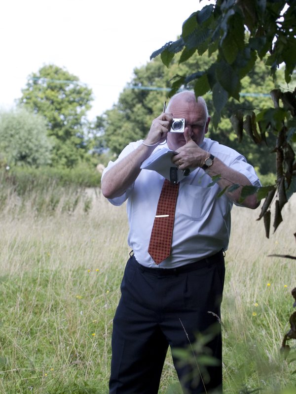 A council officer standing in long grass photographing Lucies Farm, camera raised to his eye