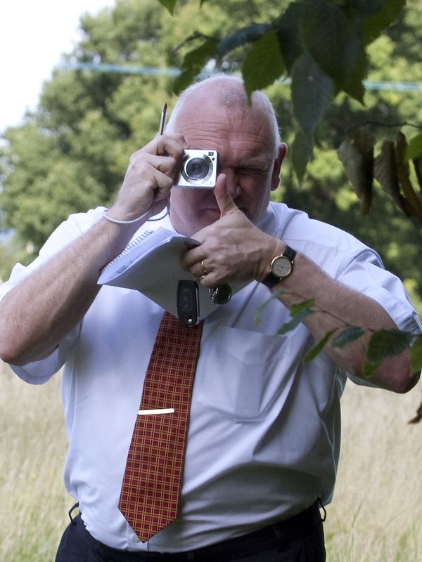 The same officer, closer: compact camera raised, notepad in his other hand