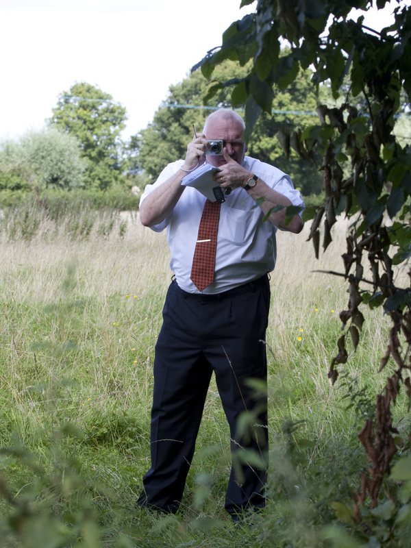 The officer raising the camera again, framed by the hedge
